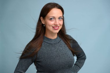 Beyaz tenli, esmer, gri kazaklı, mavi arka planda stüdyoda poz veren genç bir kadın.