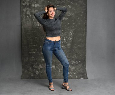 Stüdyoda gri arka planda kot pantolon giymiş gülümseyen bir kadının tam boy portresi.