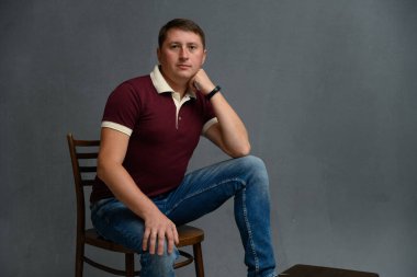 Beyaz adam gri arka planda, stüdyoda sandalyede otururken kameraya bakıyor.