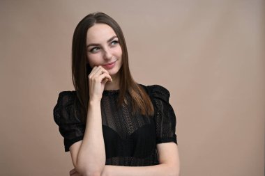 Beyaz tenli esmer, bej bir arka planda düşünceli bir gülümsemeyle yan tarafa bakıyor.