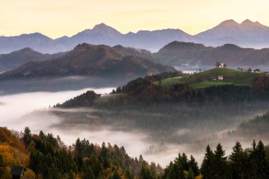 Slovenya 'nın sonbahar mevsiminin doğuşu sırasında alçak sis ve arka planda Alpler ile St. Thomas Kilisesi