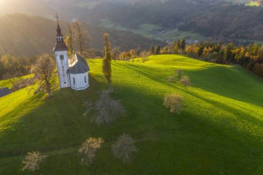 Tepelerin havadan görünüşü, renkli ormanlar ve Sv Tomaz kilisesi. Slovenya 'da sonbaharda gün batımı