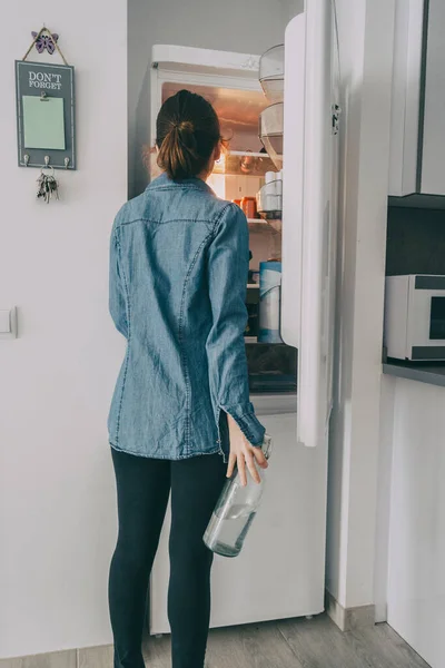 girl inside the kitchen opening the fridge door and looking inside while grabbing a bottle of water