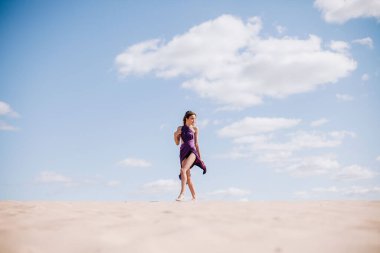 Çölde rüzgarda poz veren bej elbiseli, mor elbiseli genç bir kız.