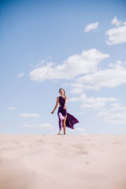 Çölde rüzgarda poz veren bej elbiseli, mor elbiseli genç bir kız.