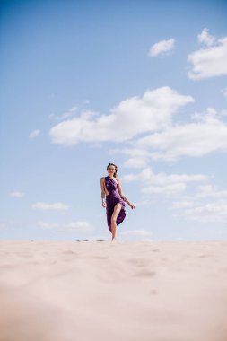 Çölde rüzgarda poz veren bej elbiseli, mor elbiseli genç bir kız.