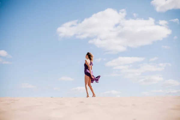 Mladá štíhlá dívka v béžových šatech s purpurovým hadříkem v rukou pózujících v poušti ve větru — Stock fotografie