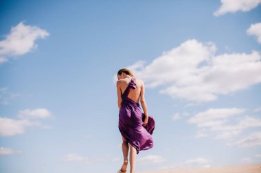 Çölde rüzgarda poz veren bej elbiseli, mor elbiseli genç bir kız.