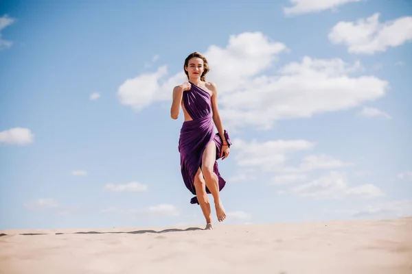 Une jeune fille mince dans une robe beige avec un tissu violet dans ses mains posant dans le désert dans le vent — Photo