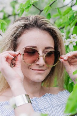 Güneş gözlüklü genç sarışın çiçek açan bir ağacın yapraklarında poz veriyor.