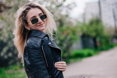 Deri ceketli ve güneş gözlüklü genç sarışın sokakta poz veriyor.