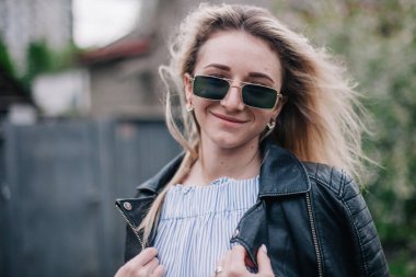 Deri ceketli ve güneş gözlüklü genç sarışın sokakta poz veriyor.