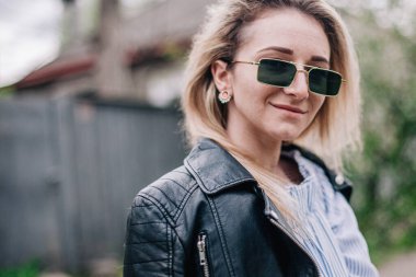Deri ceketli ve güneş gözlüklü genç sarışın sokakta poz veriyor.