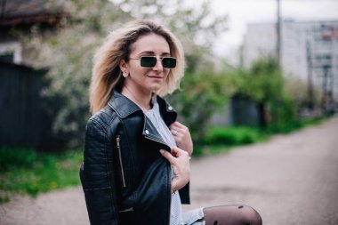 Deri ceketli ve güneş gözlüklü genç sarışın sokakta poz veriyor.