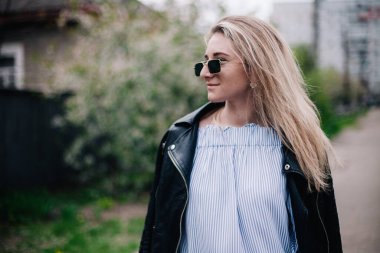 Deri ceketli ve güneş gözlüklü genç sarışın sokakta poz veriyor.