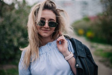 Deri ceketli ve güneş gözlüklü genç sarışın sokakta poz veriyor.