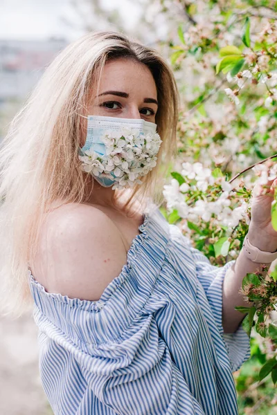 A girl in a medical mask decorated with live flowers poses in flowering trees — Foto Stock