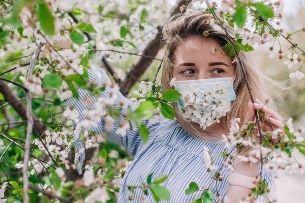 A girl in a medical mask decorated with live flowers poses in flowering trees — Stockfoto