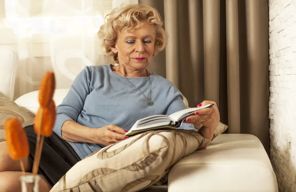 Senior mulher lendo um livro, na sala de estar — Fotografia de Stock