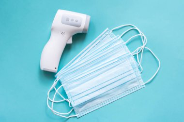 Mavi arka planda tıbbi maske yığını olan dijital kızılötesi termometre. Virüslere, gribe ve insan vücut ısısının kontrol edilmesine karşı sağlık koruma araçları