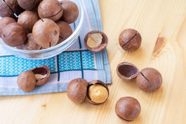 Çekirdeklerin içinde macadamia fındığı yığını ve yarısı mavi peçete ve şeffaf kâseyle ahşap arka planda açılmış. Avustralya fıstıklarının tohumları, aynı zamanda Macadamia olarak da bilinir. Makro yiyecek fotoğrafı. Yakından bak..