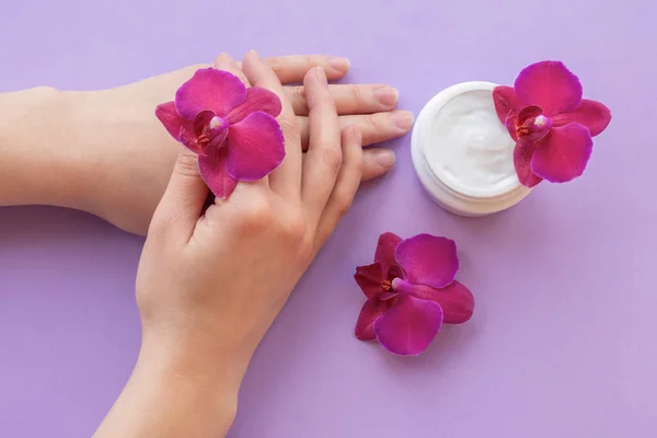 Cosmetic background with hands, white cream and fragile magenta colored orchids.Moisturizing facial cream in a jar and blossoming orchid flowers on purple background, skin care cosmetics.