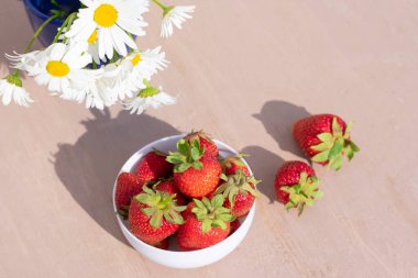 Tahta arka planda beyaz bir kasede uzun gölgeli tatlı kırmızı olgun çilekler.