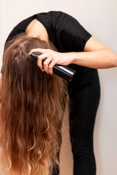 Young woman hand holds bottle with cosmetic oil and spraying her clean wavy blond long hair. Hair protection and treatment for hair loss.