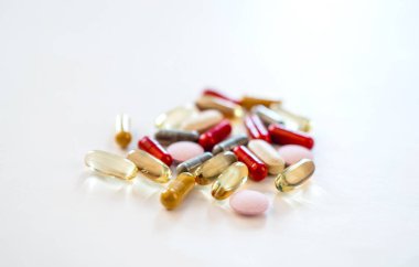 Değişik renkli kapsüller ve tabletler temiz bir tıbbi hayat sahnesinde beyaz bir zemin üzerinde açık bir cam kutudan yayılmış..