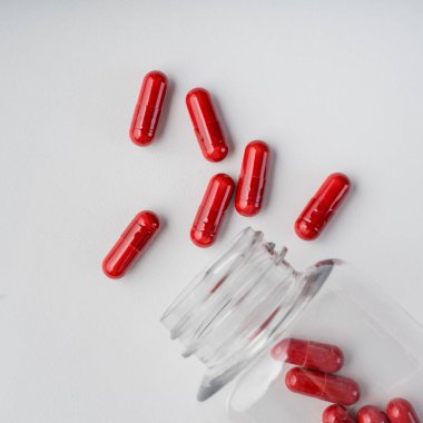 Kırmızı kapsüller beyaz arka planda açık plastik bir kutudan dökülmüş. Temiz bir tıbbi hayat olarak fotoğraflanmış..
