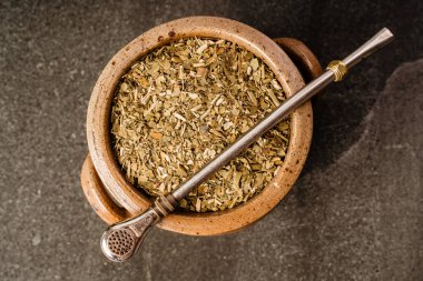 Kurumuş yerba mat, sade bir yaşam kompozisyonu içinde koyu dokulu bir arka plana yerleştirilmiş metal bombilla ile kırsal seramik bir kap içinde bırakır..