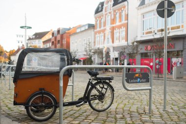 Ahşap kutulu bir kargo bisikleti, kaldırım taşı meydanında belirlenmiş bir bisiklet alanına park edilmiştir. Arka plandaki tarihi binalar, Avrupa 'daki bir şehir merkezinde çevre dostu bir şehir yaşam tarzını ön plana çıkarıyor.