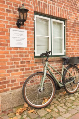 Büyük tekerli yeşil bir bisiklet beyaz çerçeveli bir pencerenin önündeki kırmızı tuğladan bir duvara park edilir. Eski bir fener küçük beyaz bir plaketin üzerine asılır. Zemin kaldırım taşlarıyla kaplı..