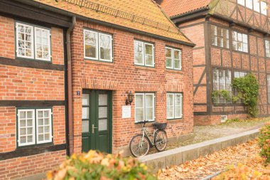 Büyük tekerli yeşil bir bisiklet beyaz çerçeveli bir pencerenin önündeki kırmızı tuğladan bir duvara park edilir. Eski bir fener küçük beyaz bir plaketin üzerine asılır. Zemin kaldırım taşlarıyla kaplı..