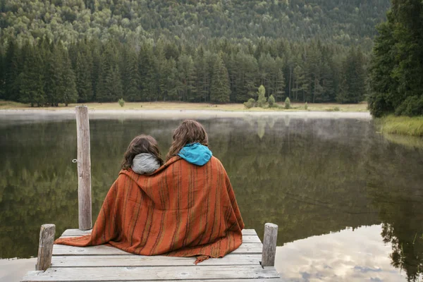 Mor och dotter sitter på en brygga med sjö och skog i f — Stockfoto