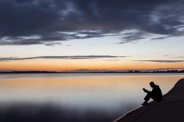 Силуэт человека, читающего у озера на закате — стоковое фото