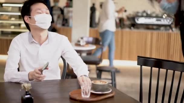 スマホを使った外科用マスクをしたアジア人男性とコーヒーショップでのオーダードリンク — ストック動画
