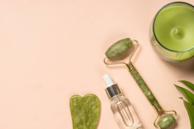 Kozmetik ürünleri - Yeşim silindirli ve kremalı gua sha masajcısı ve pastel arka planda serum şişeleri.