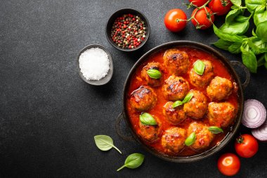 Köfteler domates sosunda, demir tabakta, siyah zemin üzerinde, yemek pişirmek için malzemelerle. Metin için boşluk olan üst görünüm.