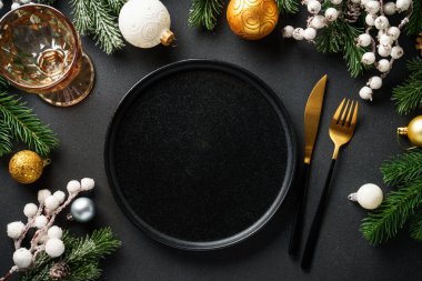 Noel sofrası. El işi tabağı, şarap kadehi, çatal bıçak takımı ve siyah arka planda altın Noel süslemeleri. Düz yatıyordu.