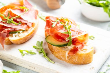 Beyaz arka planda kesme tahtasının üzerinde prosciutto ve roka ile sandviç açın. Resmi kapat.