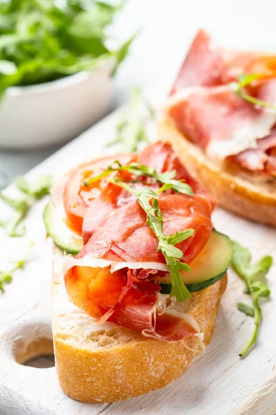 Beyaz arka planda kesme tahtasının üzerinde prosciutto ve roka ile sandviç açın. Resmi kapat.