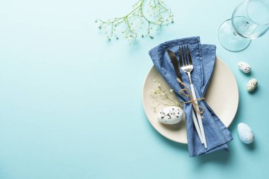 Mavi üzerine Paskalya masası. Boş tabak, çatal bıçak takımı, paskalya yumurtası ve çiçekler. Düz görünüm.