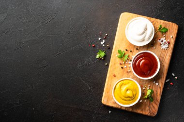 Kesme tahtasında ketçap, mayonez ve hardal var. Üst görünüm, tasarım alanı, koyu arkaplan.