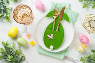 Paskalya kompozisyonu. Tatil masası süslemeleri, beyaz tabak, altın çatal bıçak, cam ve renkli paskalya yumurtaları. Üst görünüm.