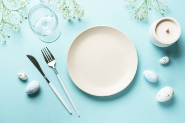 Mavi üzerine Paskalya masası. Boş tabak, çatal bıçak takımı, paskalya yumurtası ve çiçekler. Düz görünüm.