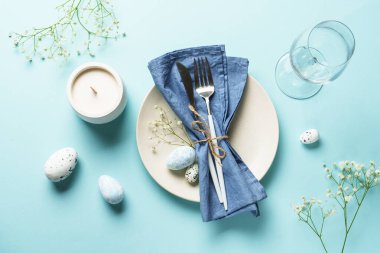 Mavi üzerine Paskalya masası. Beyaz tabak, çatal bıçak, Paskalya yumurtası ve çiçekler..