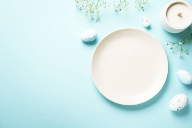 Mavi üzerine Paskalya masası. Boş tabak, çatal bıçak takımı, paskalya yumurtası ve çiçekler. Düz görünüm.
