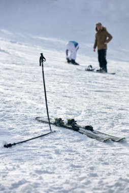 Bir çift kayak ve sopa karda sıkışmış, insanlar arka planda.