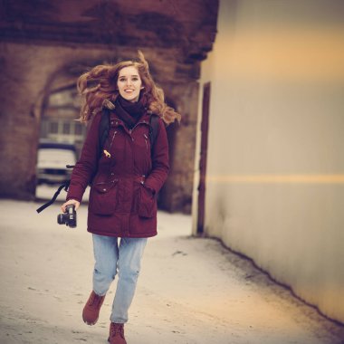 Kış caddesinde yürüyen kameralı mutlu kadın fotoğrafçı, sıcak, klasik tonlu bir görüntü.